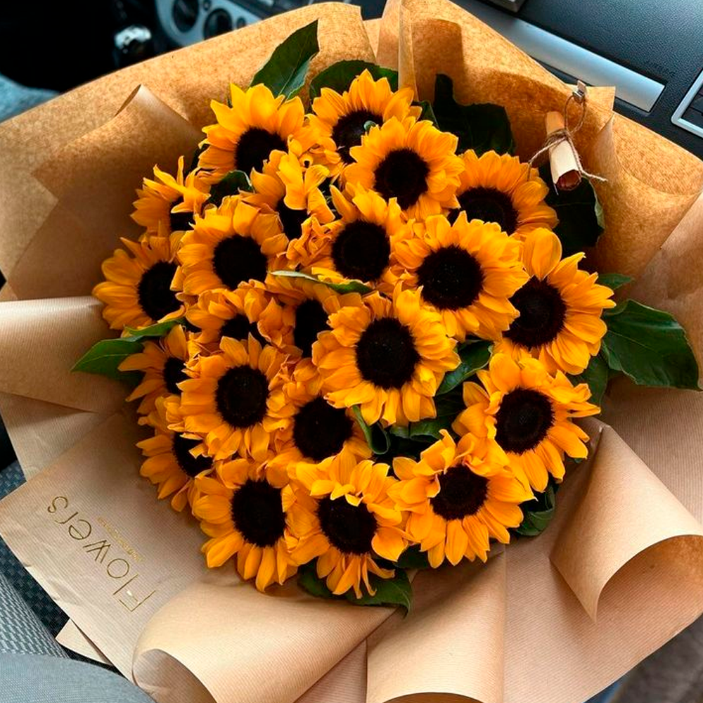 A bouquet of bright yellow sunflowers with dark centers is wrapped in brown paper, placed on a car seat. A small paper tag reading "FLOWERS" is attached to the bouquet.