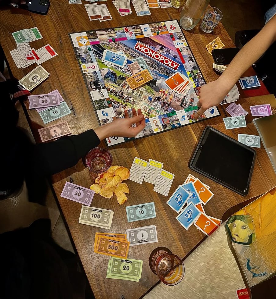 Four people play Monopoly at a wooden table cluttered with Monopoly money, cards, game pieces, snacks, drinks, and a digital tablet. Hands reach toward the board, suggesting active gameplay.