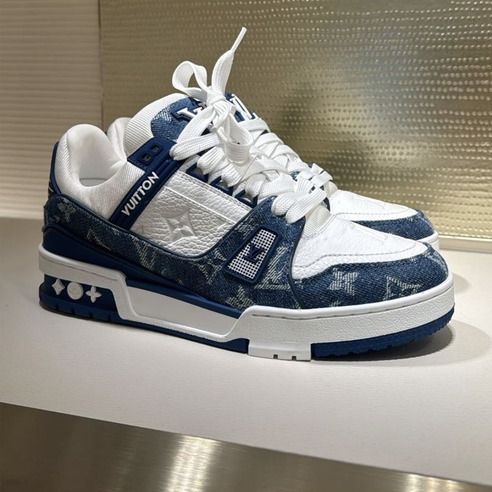 A pair of Louis Vuitton sneakers with white leather and blue denim panels featuring the LV monogram, white laces, and a blue and white rubber sole, placed on a reflective surface.