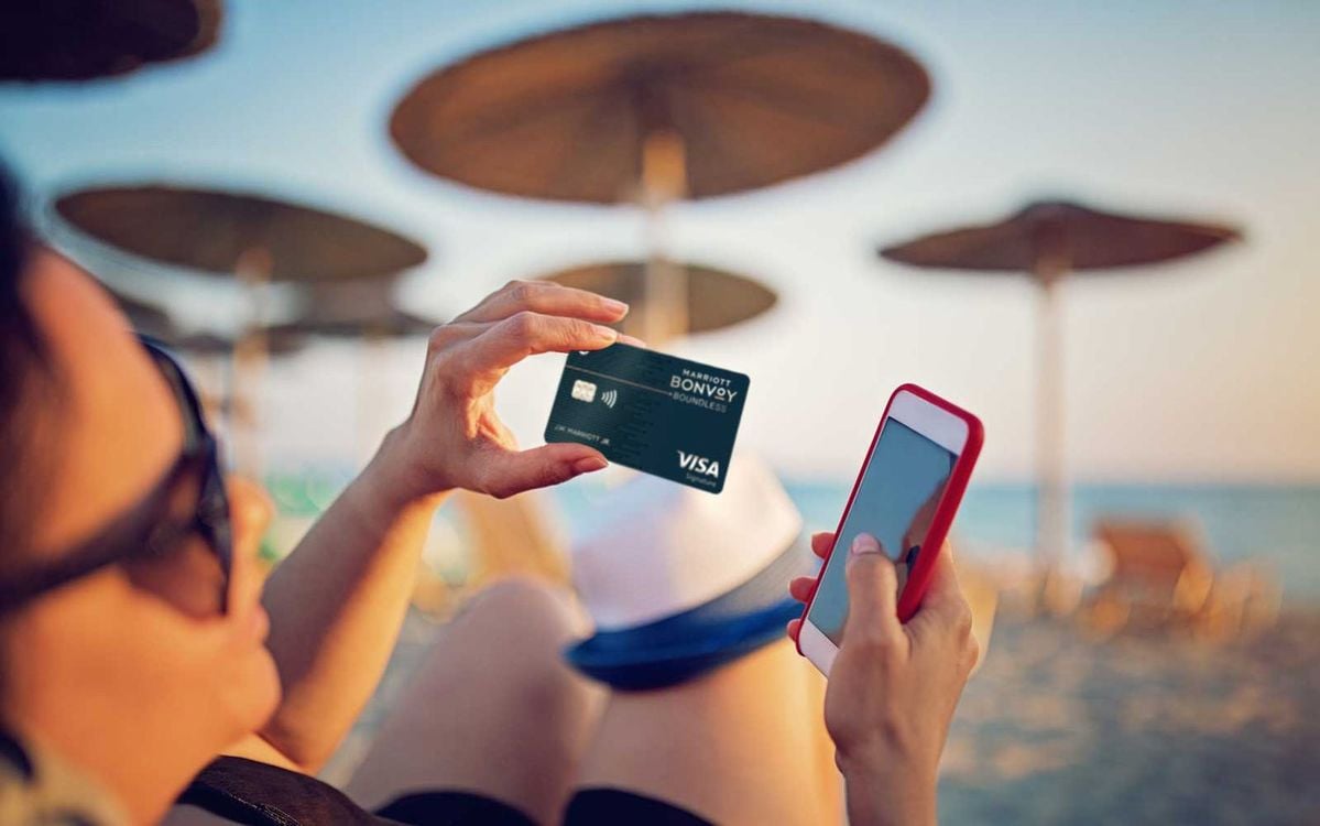 A person relaxes on a beach chair, holding a credit card in one hand and a smartphone in the other, with blurred beach umbrellas and sand in the background.