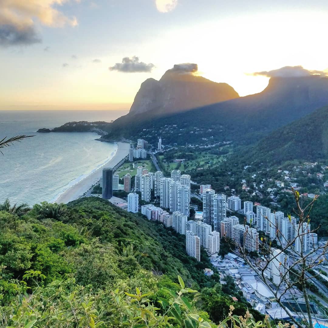 A scenic view of a coastal city with tall buildings near a sandy beach, surrounded by lush green hills and mountains at sunset. The ocean extends to the left, and the sky is partly cloudy with warm sunlight.