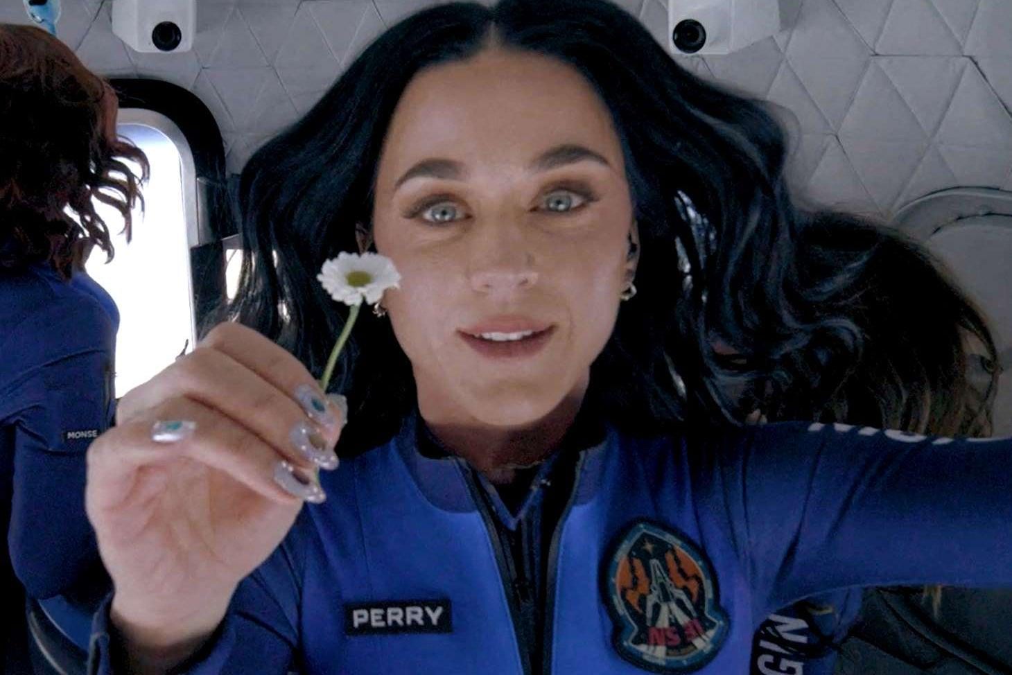 A woman with long dark hair in a blue spacesuit floats in zero gravity, holding a small white daisy. Her suit has a patch and a name tag that says "PERRY." The background suggests the inside of a spacecraft.
