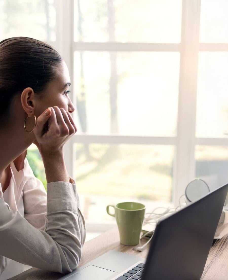 A woman sits at a desk with a laptop and green mug, gazing thoughtfully out a bright window, resting her chin on her hand.