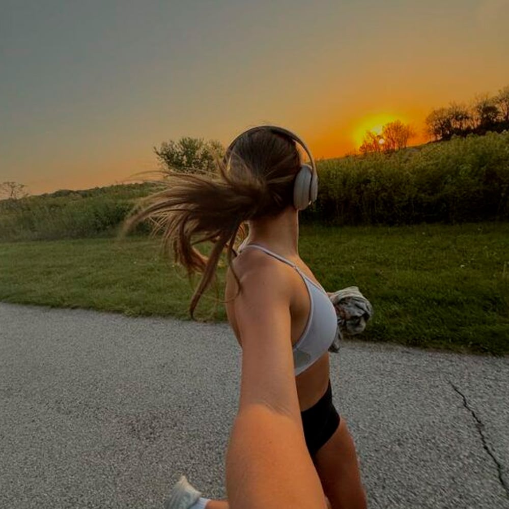 A woman wearing headphones, a sports bra, and shorts jogs on a paved path at sunset, holding a shirt in one hand and taking a selfie with the other. Trees and grass line the path under an orange sky.