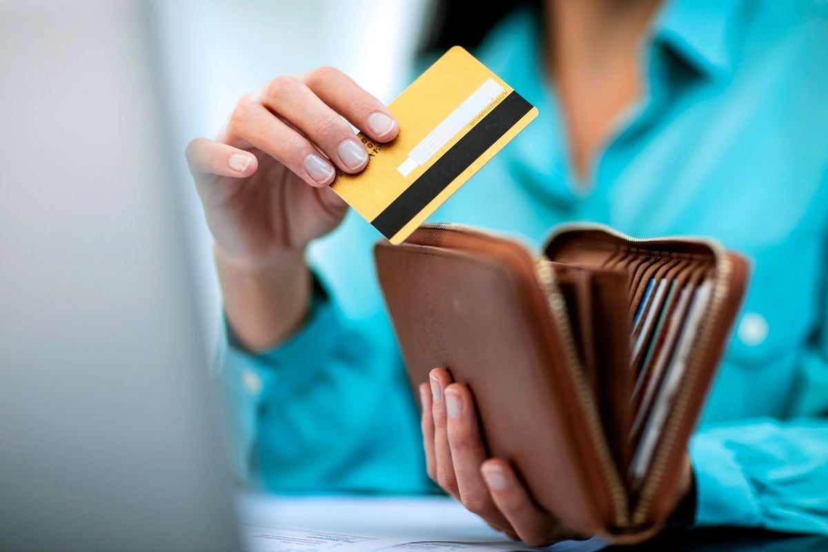 A person wearing a teal shirt is holding a brown wallet and taking out a yellow credit card. The scene is close-up, focusing on the hands and wallet. A blurred laptop is visible in the foreground.