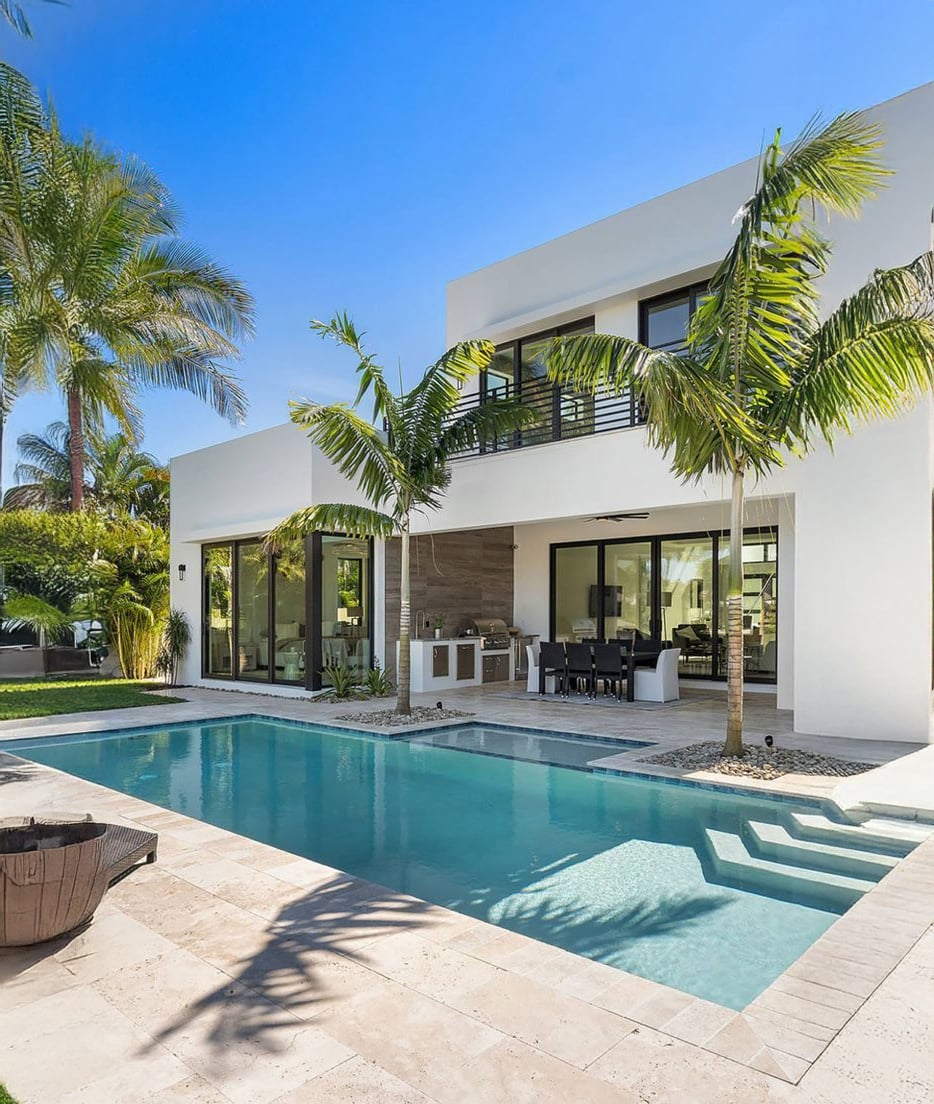 Modern white two-story house with large windows, surrounded by palm trees, features a backyard patio, outdoor dining area, and a rectangular swimming pool on a sunny day.