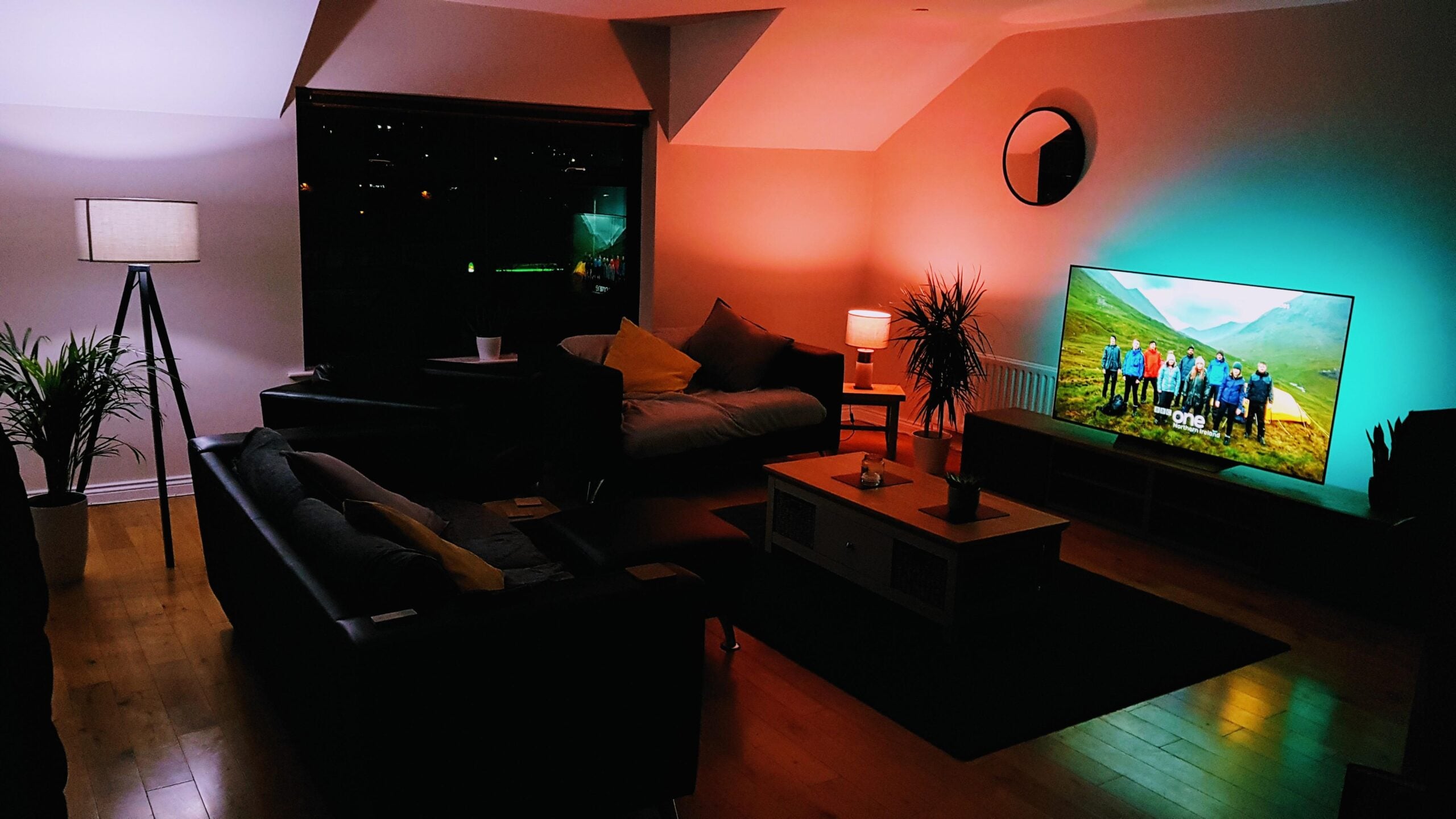 A cozy living room at night with modern furniture, several lamps, a potted plant, a coffee table, and a TV displaying a nature scene. The room is softly lit with warm and colorful ambient lighting.