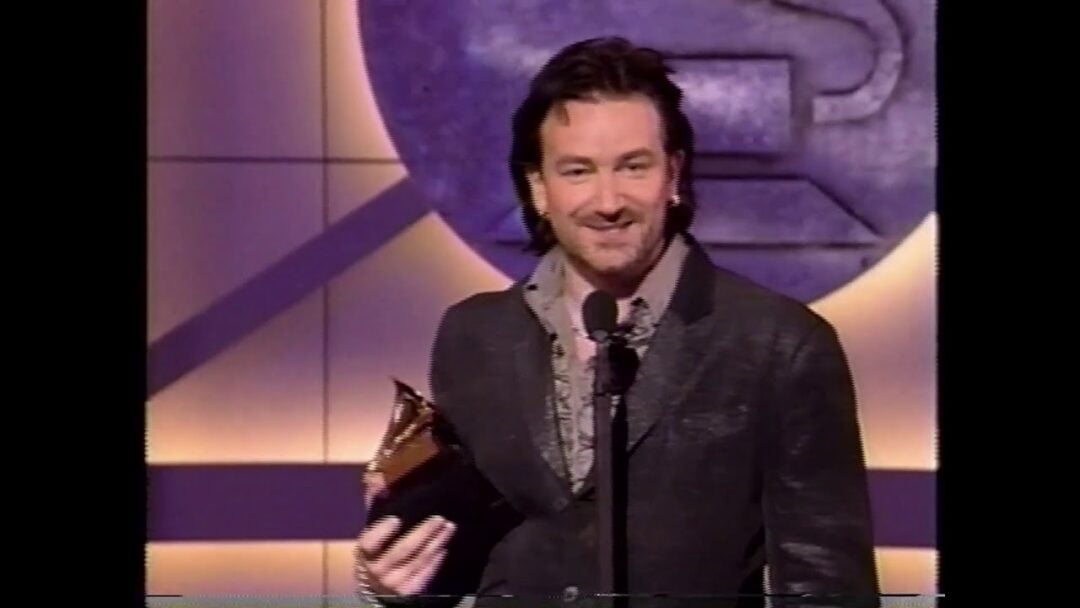A man in a suit holds a Grammy Award and smiles while standing at a microphone on stage, with a large circular emblem in the background.
