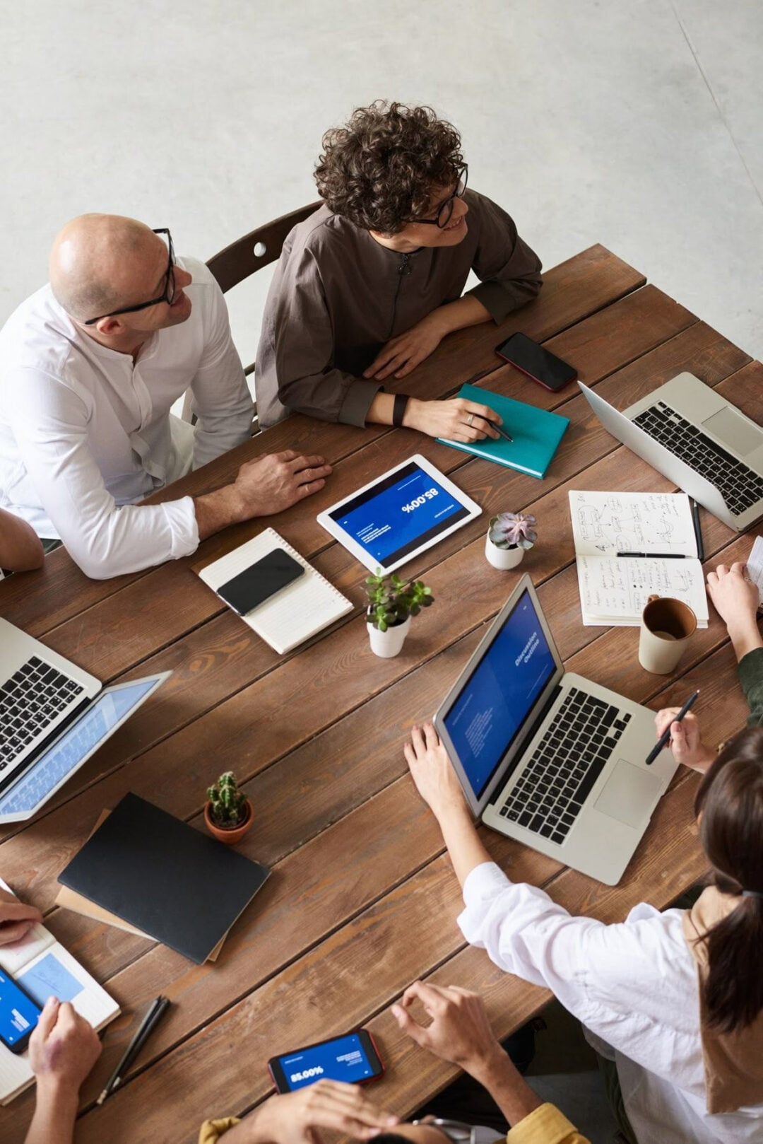 A group of people sit around a wooden table with laptops, tablets, notebooks, and coffee cups, collaborating and discussing during a meeting in a modern workspace.