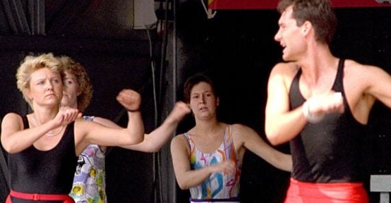 Four people in colorful workout outfits are participating in an energetic aerobics class, moving their arms rhythmically. The background is dim, highlighting their exercise movements.