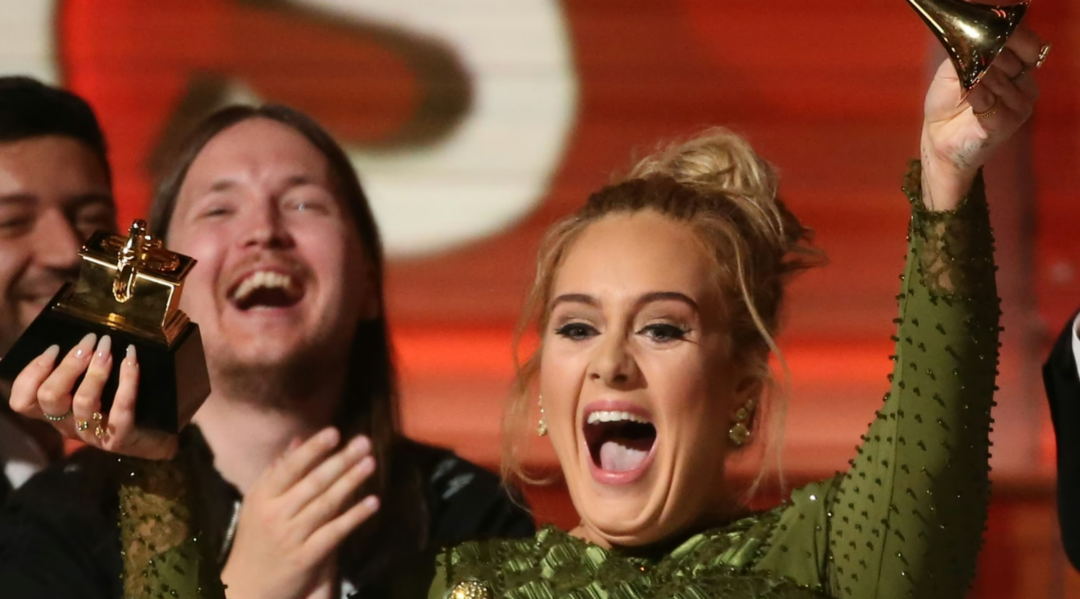 A woman in a green dress joyfully holds up a Grammy award, smiling widely with her mouth open, while a man next to her claps and laughs. The background is red and blurred.