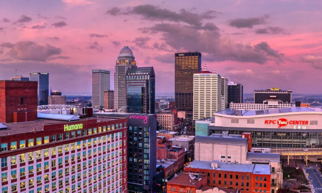 A city skyline at sunset with tall buildings, including the KFC Yum! Center and Humana offices, under a pink and purple sky with scattered clouds.