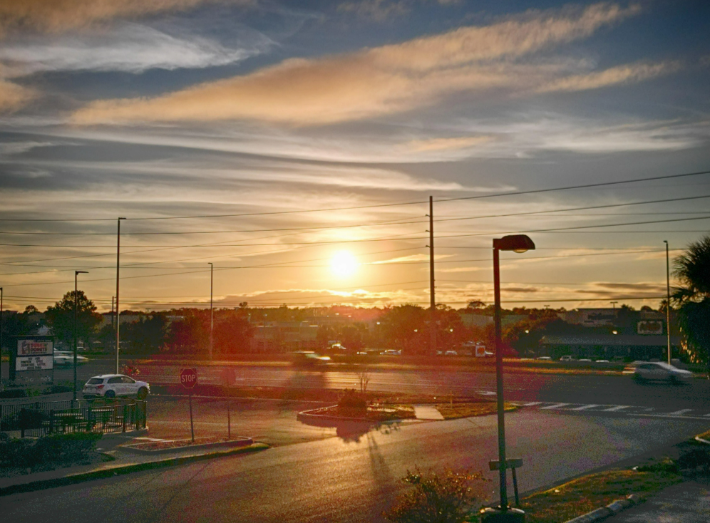A golden sunset over a busy street with cars, a stop sign, streetlights, and scattered clouds in the sky, casting warm light on the scene.