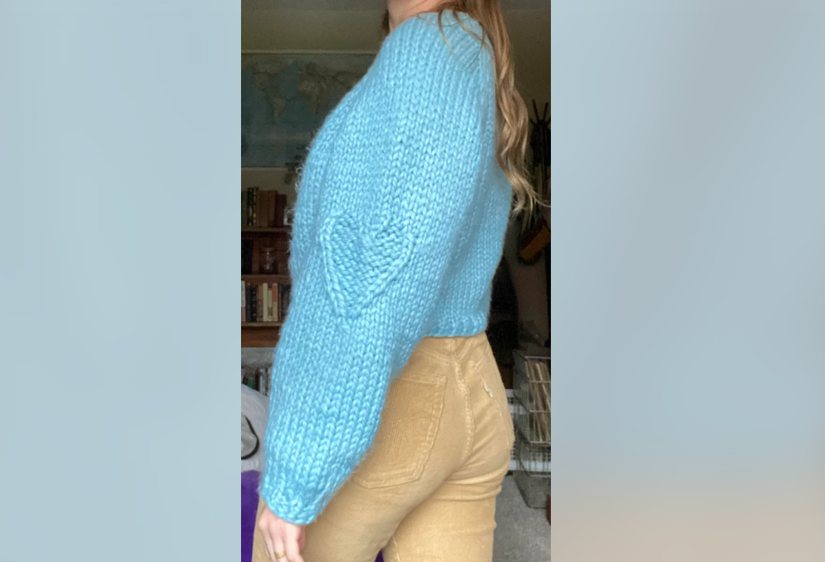 A person stands sideways indoors, wearing a chunky light blue knit sweater with a heart pattern on the sleeve and light brown pants. Bookshelves and a world map are visible in the background.