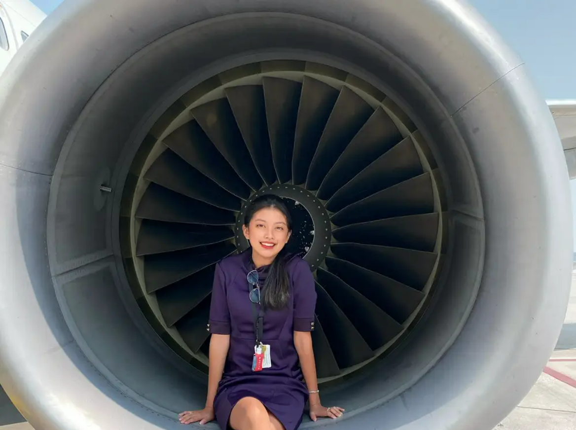 A woman in a dark uniform with a name badge sits inside the engine of an aircraft, smiling at the camera. The large fan blades of the jet engine form a circular background behind her.