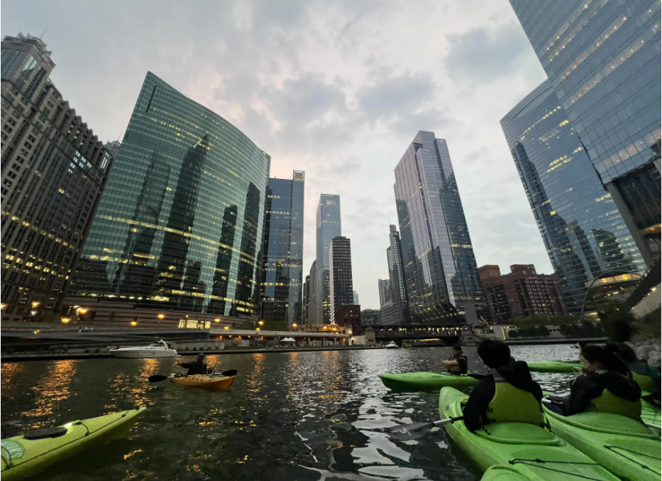 People kayaking on a river surrounded by tall, modern skyscrapers and office buildings at dusk, with lights reflecting on the water and a cloudy sky overhead.
