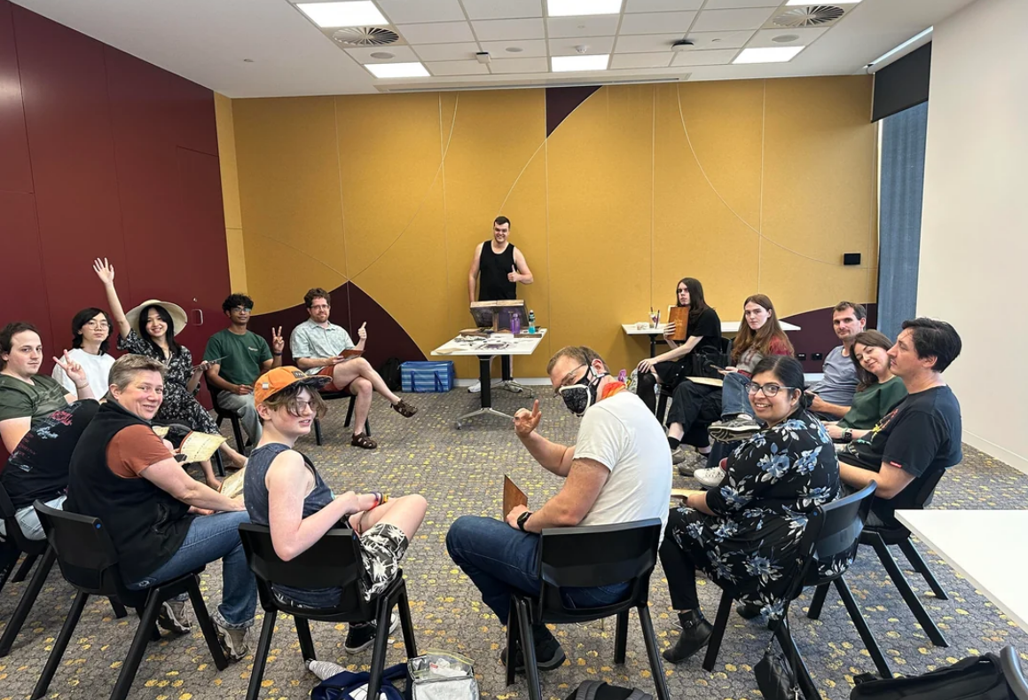 A group of people sitting in a circle in a brightly lit room, some smiling and waving at the camera. A person stands at a table in the background. The atmosphere appears friendly and relaxed.