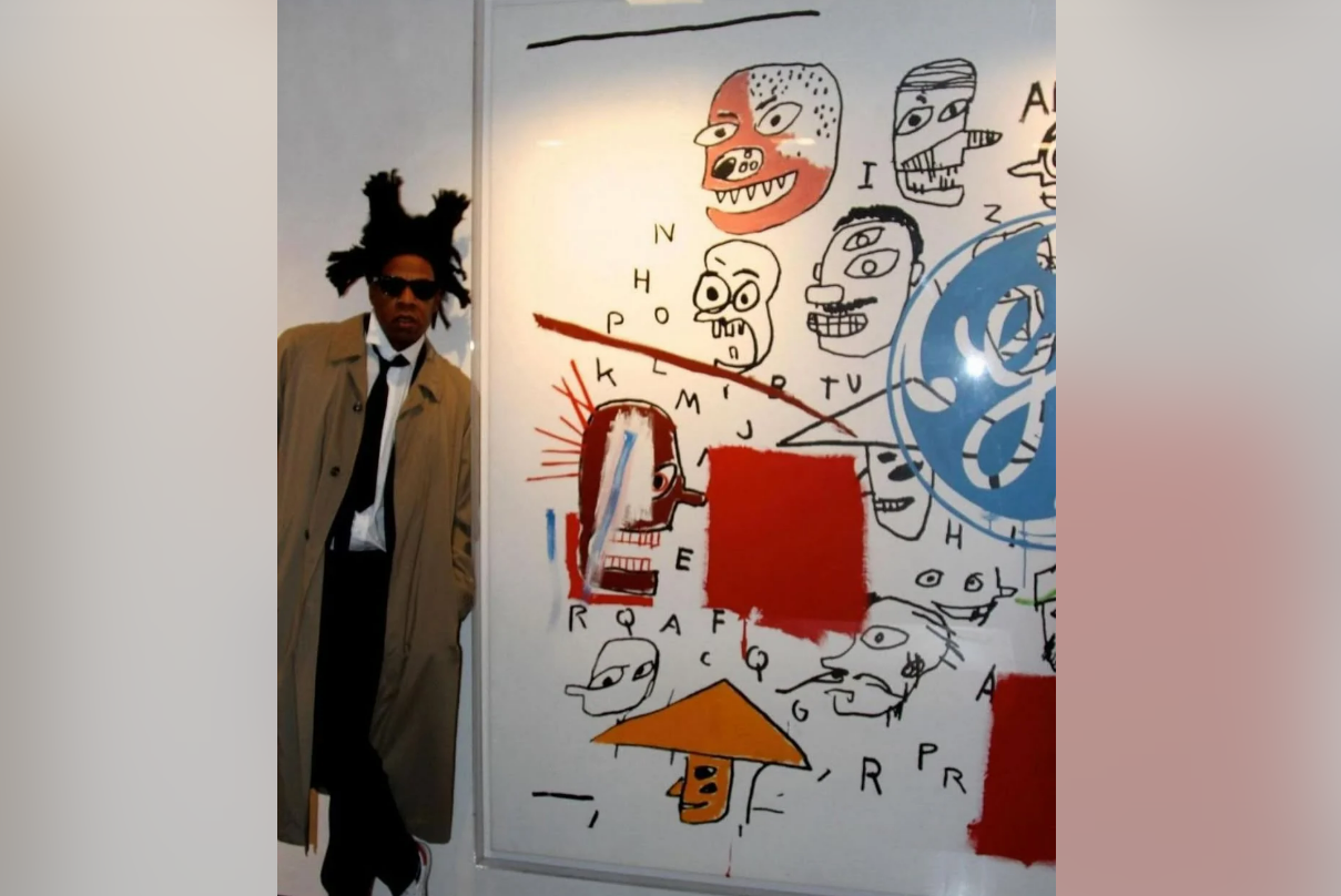 A man in a trench coat and sunglasses stands next to a large abstract artwork featuring cartoonish faces, letters, numbers, red rectangles, and blue shapes on a white background.