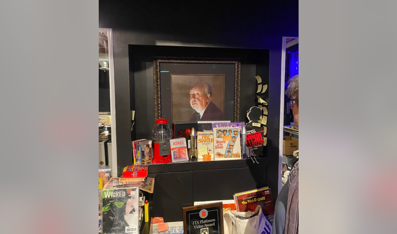 A framed portrait of an older man is displayed on a shelf surrounded by memorabilia, including comic books, DVDs, a red Atari container, and various collectibles. Part of a person is visible on the right edge of the image.
