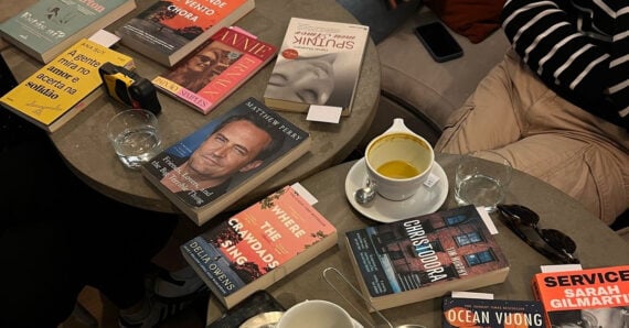 Several books are arranged on two round tables, alongside a cup of tea, a phone, and some personal items. Two people are sitting nearby, one wearing black pants, the other in black and white stripes.