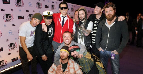 A group of ten men, some dressed casually and others in costumes, pose together on a purple carpet at an MTV EMA event, with event branding and logos visible in the background.