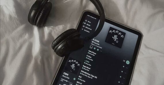 A pair of black headphones rests on a white bed next to a tablet displaying a music playlist with a dark-themed interface.