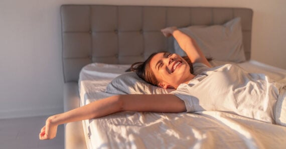 A woman is lying in bed, smiling and stretching her arms out wide. Morning sunlight shines on her, giving the room a warm and peaceful atmosphere.