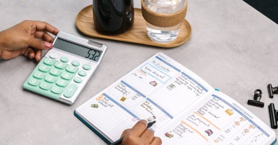 A person uses a large calculator and writes in a colorful budget planner notebook on a gray table, with a glass of water, a dark bottle on a wooden tray, and binder clips nearby.