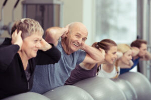 A group of adults, both men and women, exercise indoors using stability balls, smiling and holding their hands behind their heads in a row.