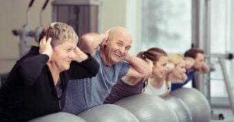 A group of adults, both men and women, exercise indoors using stability balls, smiling and holding their hands behind their heads in a row.