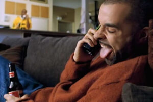 A man in a red sweater sits on a couch, holding a beer bottle and talking on the phone with his tongue out. Another person stands in the background, near a kitchen counter.