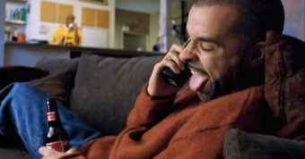 A man in a red sweater sits on a couch, holding a beer bottle and talking on the phone with his tongue out. Another person stands in the background, near a kitchen counter.