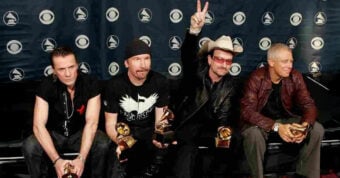Four men sit on a bench holding Grammy Awards in front of a step-and-repeat backdrop. One man wears a cowboy hat and sunglasses, raising a peace sign, while the others hold their awards and face the camera.