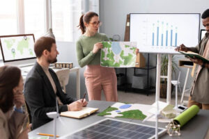 Four people in an office discuss environmental maps and data; one woman presents a map, a man writes in a notebook, and charts, maps, and solar panels are visible on tables and monitors around them.