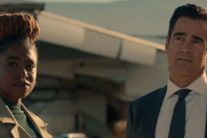 A woman with braided hair and a man in a suit stand outdoors in front of a blurred airplane, both looking slightly to the side with serious expressions.