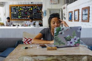 A woman sits at a wooden table with a floral-laptop in a cozy café, wearing headphones and smiling. The café has a thatched roof, menu board, framed pictures, and a "No Smoking" sign on the wall.