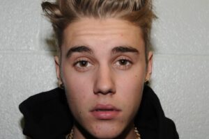 A young person with short, styled blonde-brown hair and light brown eyes looks directly at the camera. They are wearing a black hoodie and gold earrings, with a neutral facial expression against a light-colored textured wall.