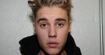 A young person with short, styled blonde-brown hair and light brown eyes looks directly at the camera. They are wearing a black hoodie and gold earrings, with a neutral facial expression against a light-colored textured wall.