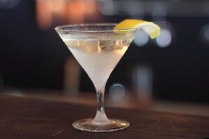 A classic martini cocktail in a clear martini glass, garnished with a twist of lemon peel, sits on a dark wooden bar with a blurred background.
