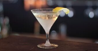 A classic martini cocktail in a clear martini glass, garnished with a twist of lemon peel, sits on a dark wooden bar with a blurred background.