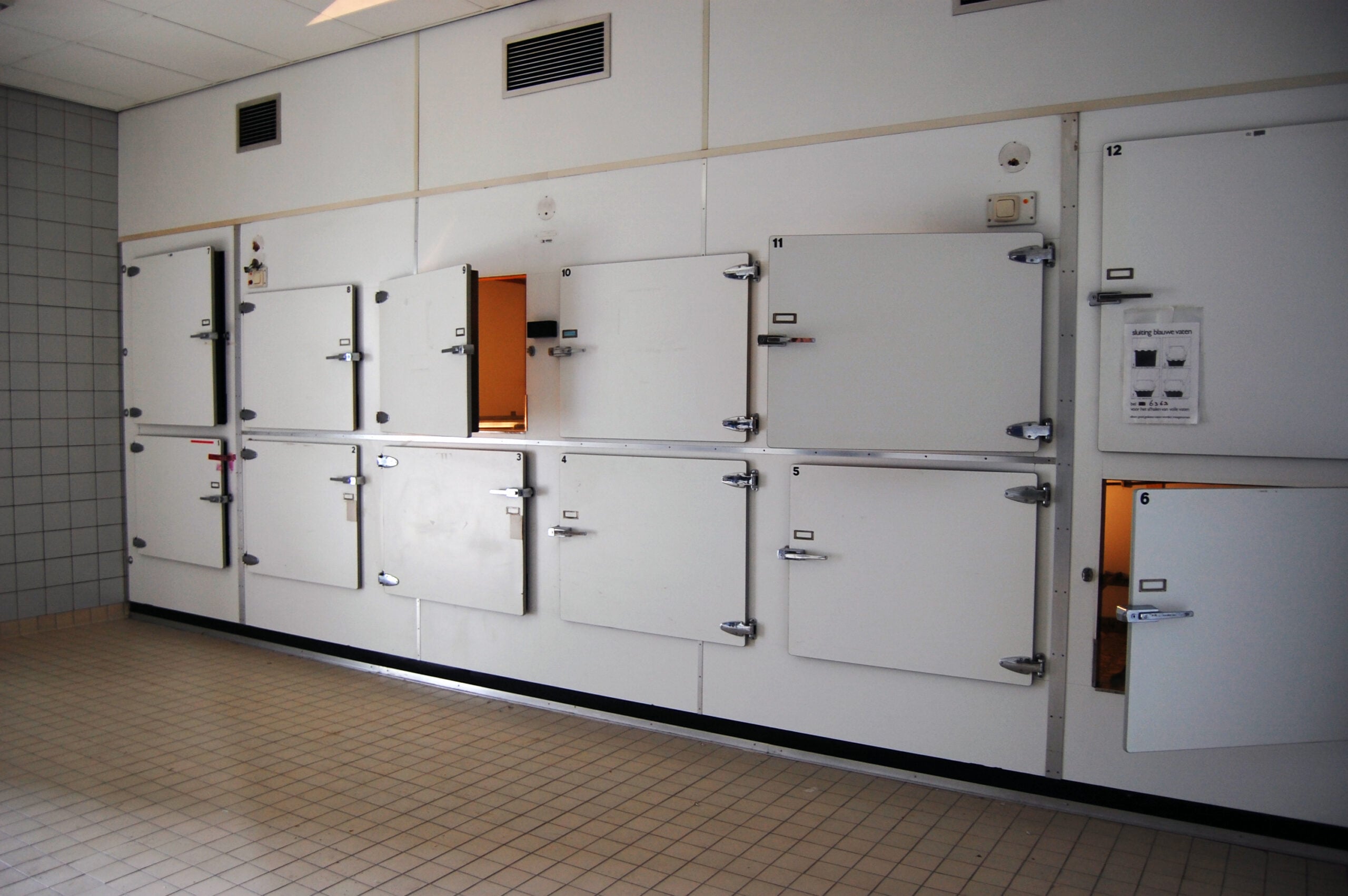 A row of white mortuary cold storage compartments with several doors partially open, located in a tiled room with beige flooring and a tiled wall to the left.