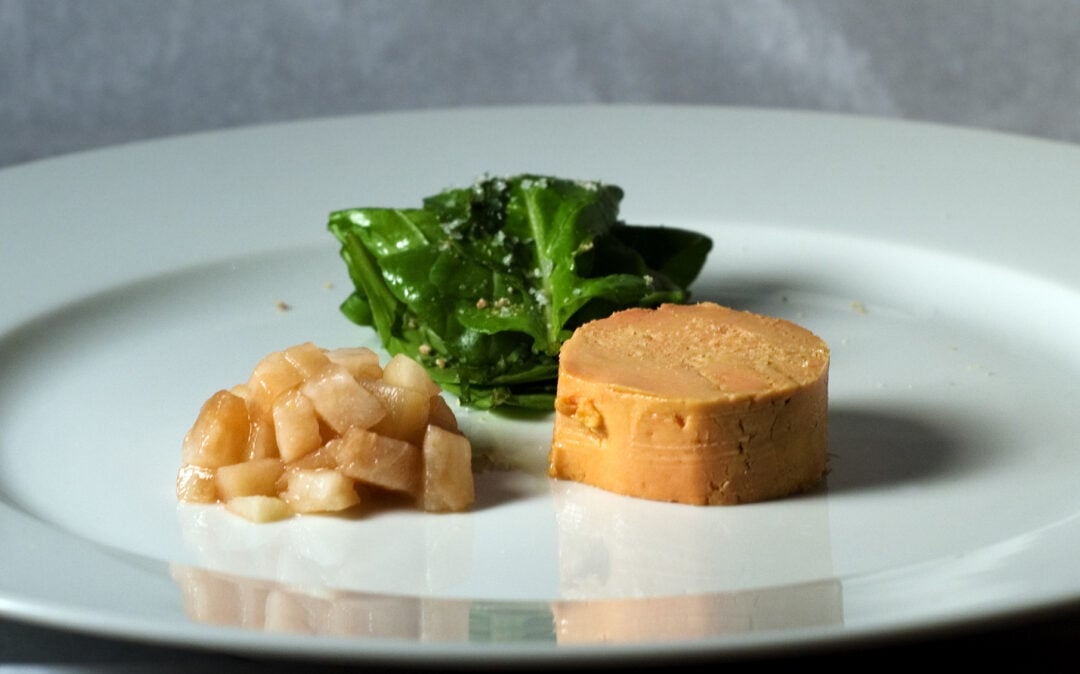 A plated dish featuring a round slice of foie gras, a small mound of diced pears, and a small pile of leafy greens on a white plate.