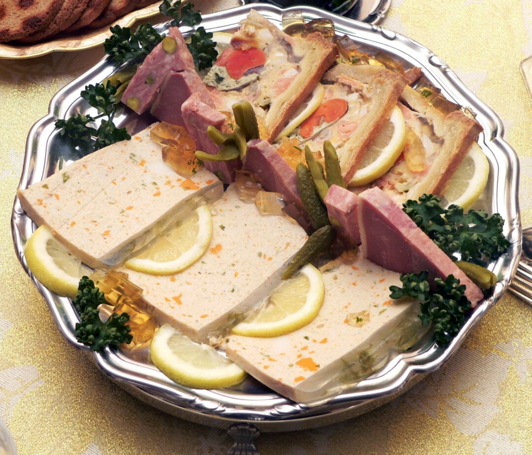 A silver platter with assorted pâté, terrine slices, and meat pâté en croûte, garnished with lemon slices, parsley, small pickles, and clear aspic cubes on a patterned tablecloth.