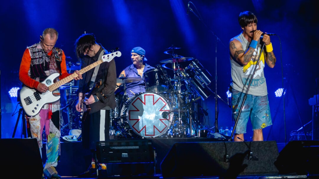 Four members of a rock band perform on stage at night; the bassist and guitarist play on the left, the drummer is centered behind a drum kit with a red star logo, and the vocalist sings into a microphone on the right.