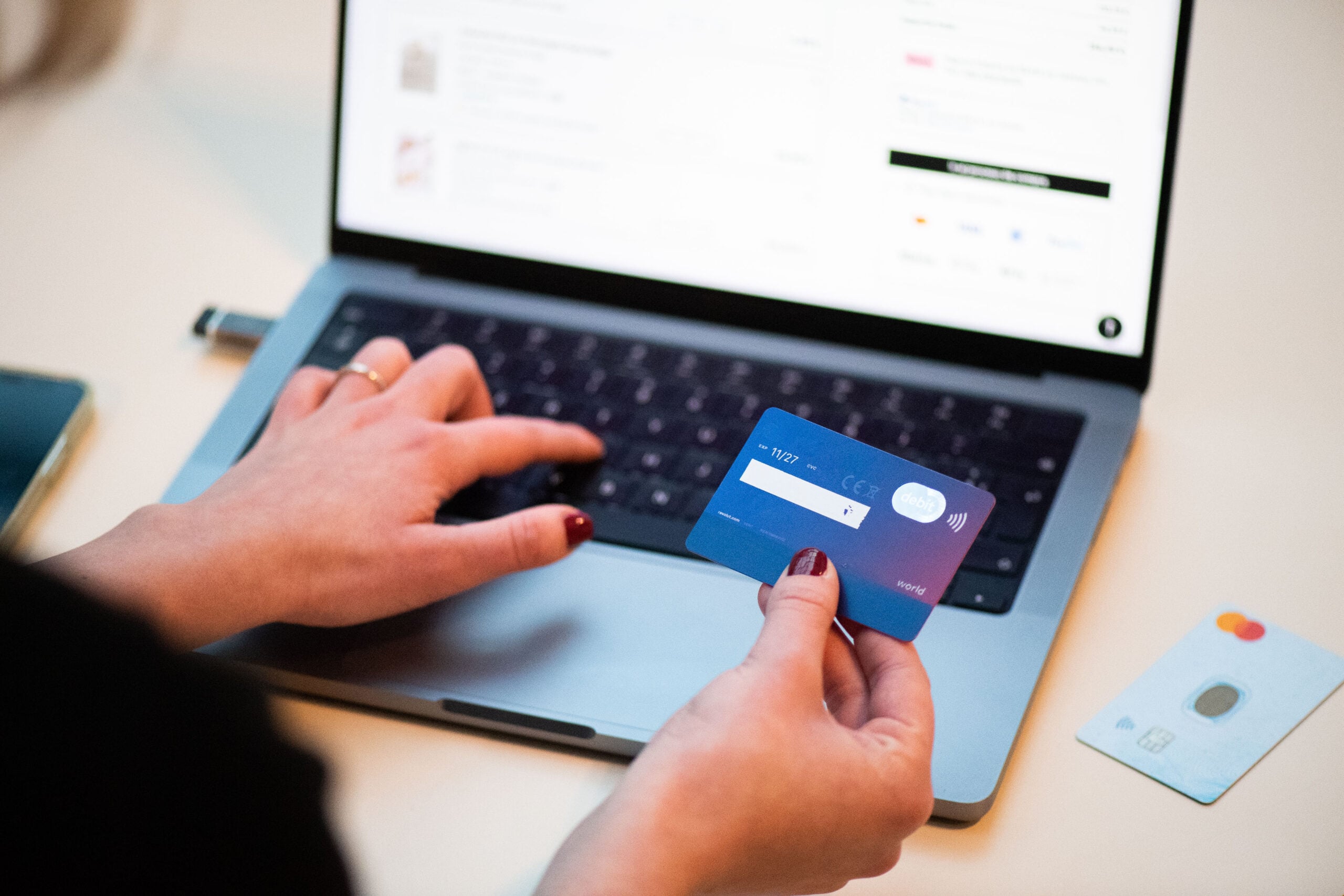 A person holds a blue credit card in one hand and types on a laptop keyboard with the other, suggesting online shopping or payment. Another card lies on the table next to the laptop.