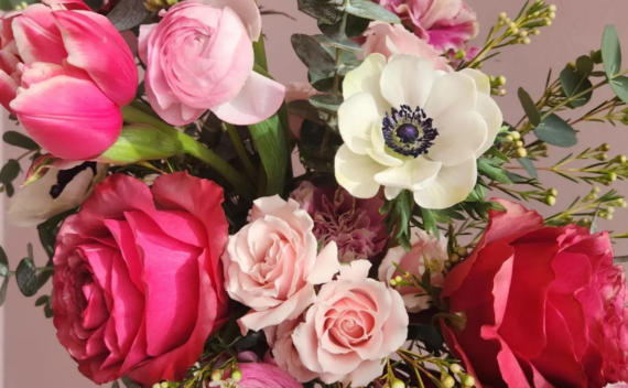 A close-up of a flower bouquet with pink roses, tulips, and a white anemone, surrounded by green leaves and small buds against a soft pink background. Valentines.