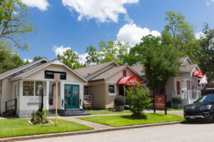 A row of quaint small shops with colorful awnings and well-kept lawns lines a sunny street. A black SUV is parked at the curb, and leafy trees stand in the background under a partly cloudy blue sky.