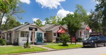 A row of quaint small shops with colorful awnings and well-kept lawns lines a sunny street. A black SUV is parked at the curb, and leafy trees stand in the background under a partly cloudy blue sky.
