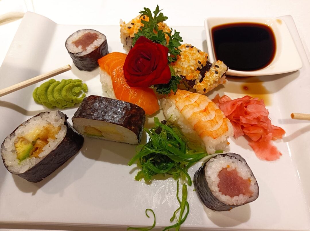 A plate of assorted sushi including rolls, nigiri with salmon and shrimp, garnished with a red rose, wasabi, pickled ginger, seaweed salad, parsley, and a dish of soy sauce. Chopsticks rest on the plate's edge.