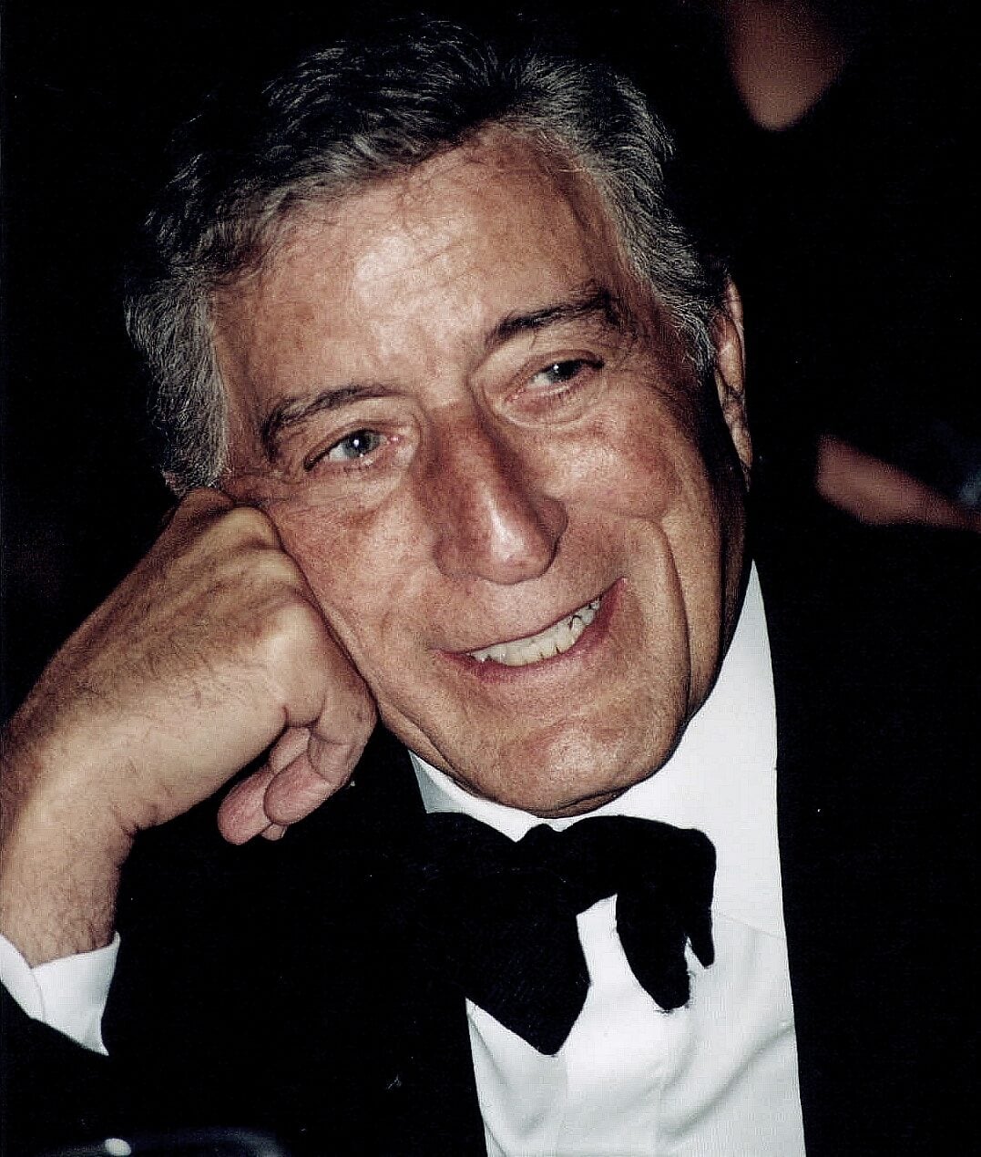 An older man with gray hair, wearing a black tuxedo and bow tie, smiles warmly while resting his head on his hand against a dark background.